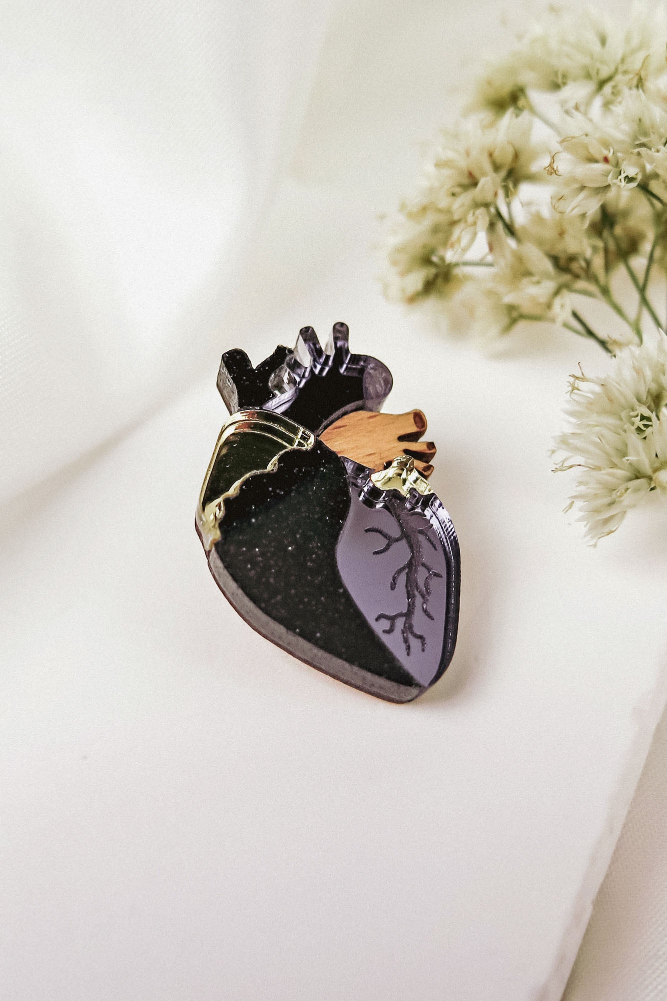 A handcrafted anatomical heart pin with black, purple, gold, and brown details rests on a white surface beside delicate white flowers—a striking statement piece for any collection.