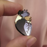 A hand holds a handcrafted Anatomical Heart Pin, shaped with metallic gold and dark accents, making it a true statement piece against the soft pink fabric background.