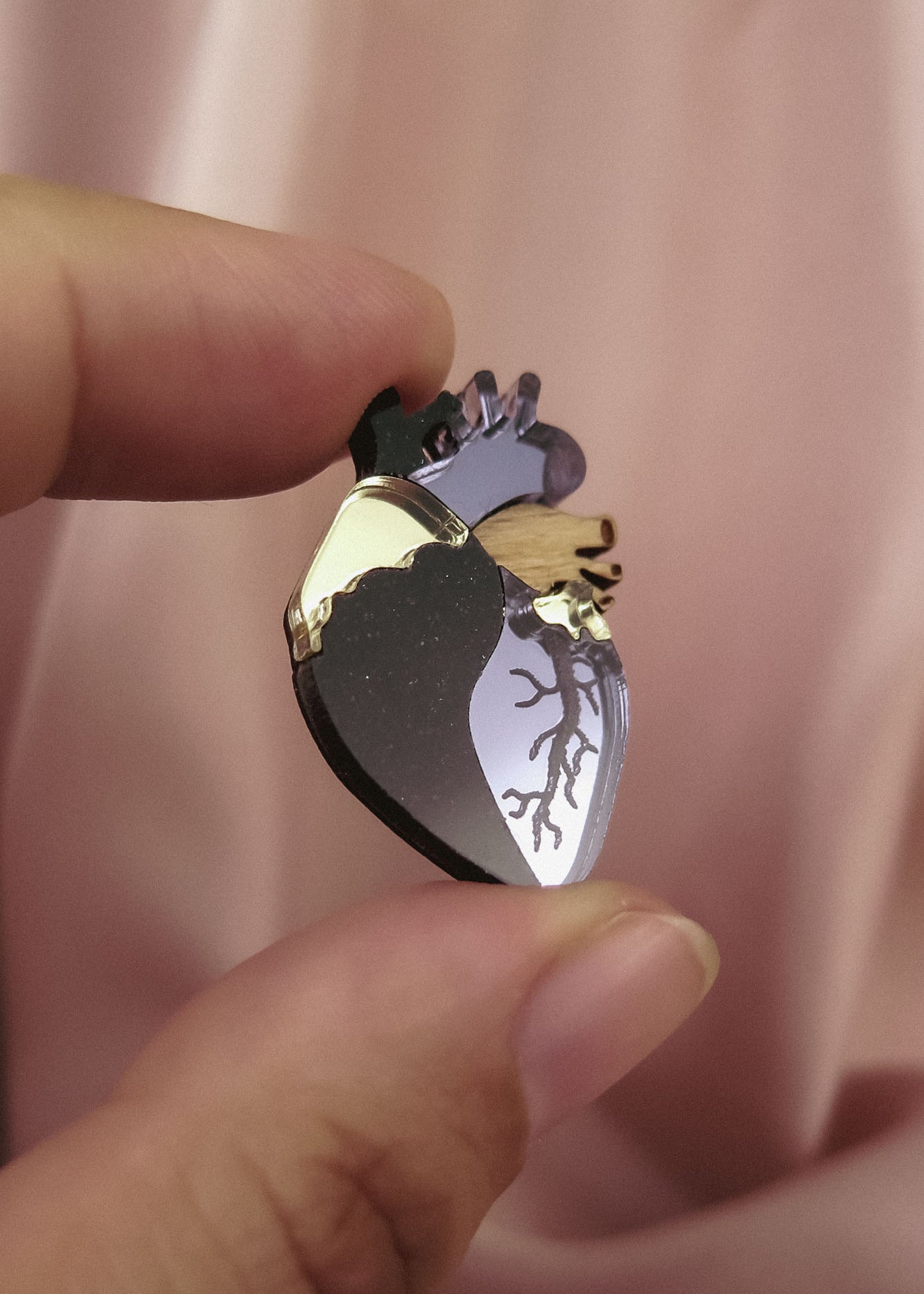 A hand holds a handcrafted Anatomical Heart Pin, shaped with metallic gold and dark accents, making it a true statement piece against the soft pink fabric background.