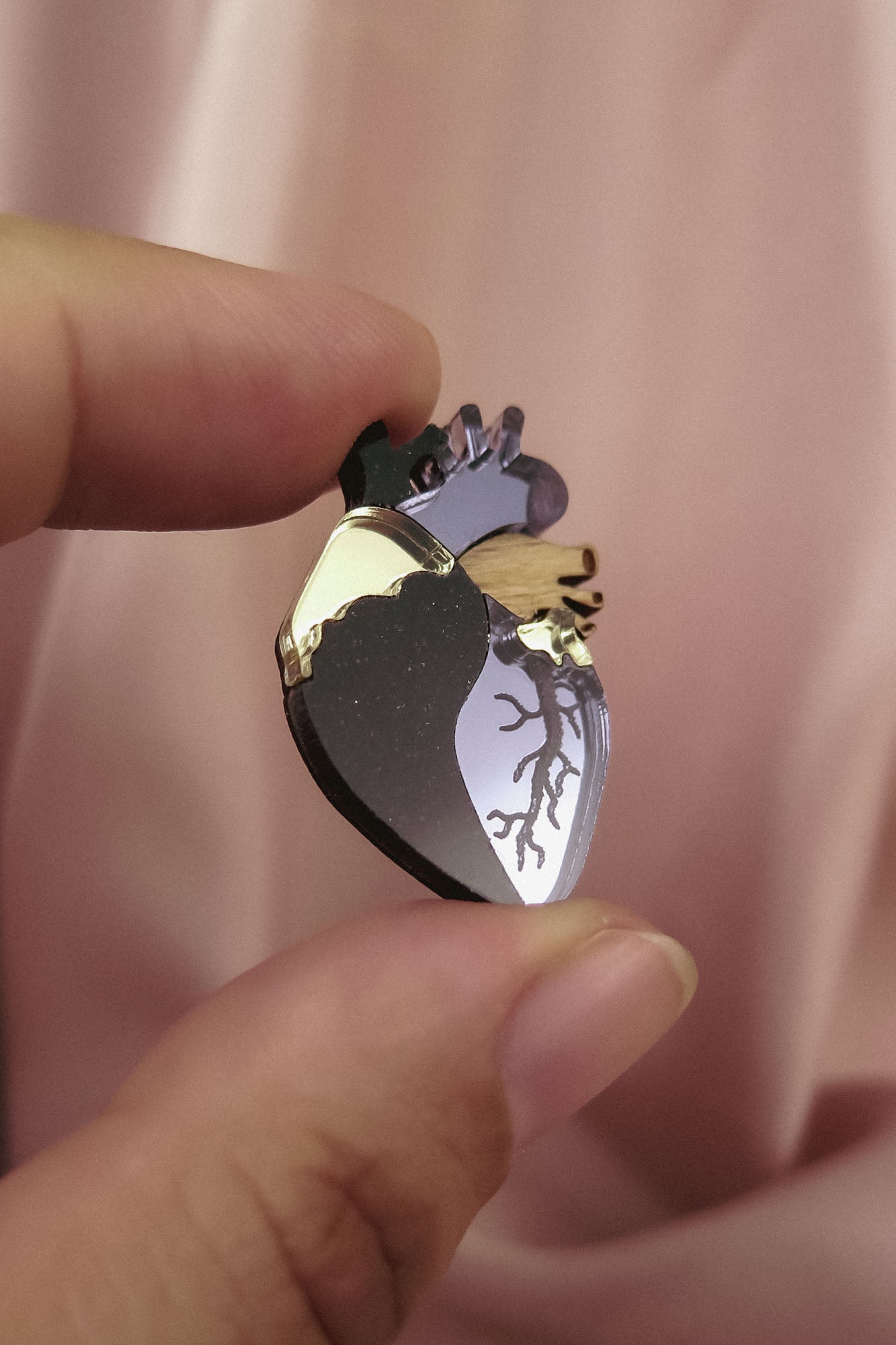 A hand holds a handcrafted Anatomical Heart Pin, shaped with metallic gold and dark accents, making it a true statement piece against the soft pink fabric background.