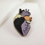 A handcrafted Anatomical Heart Pin, this decorative enamel statement piece features black, gold, and purple sections with detailed veins, beautifully displayed on a white fabric background.