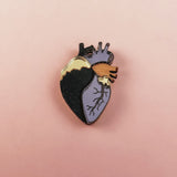 A handcrafted Anatomical Heart Pin with one side black, the other lavender, and highlighted by gold and purple accents. This statement piece rests on a pink background, perfect for adding bold flair to your collection.