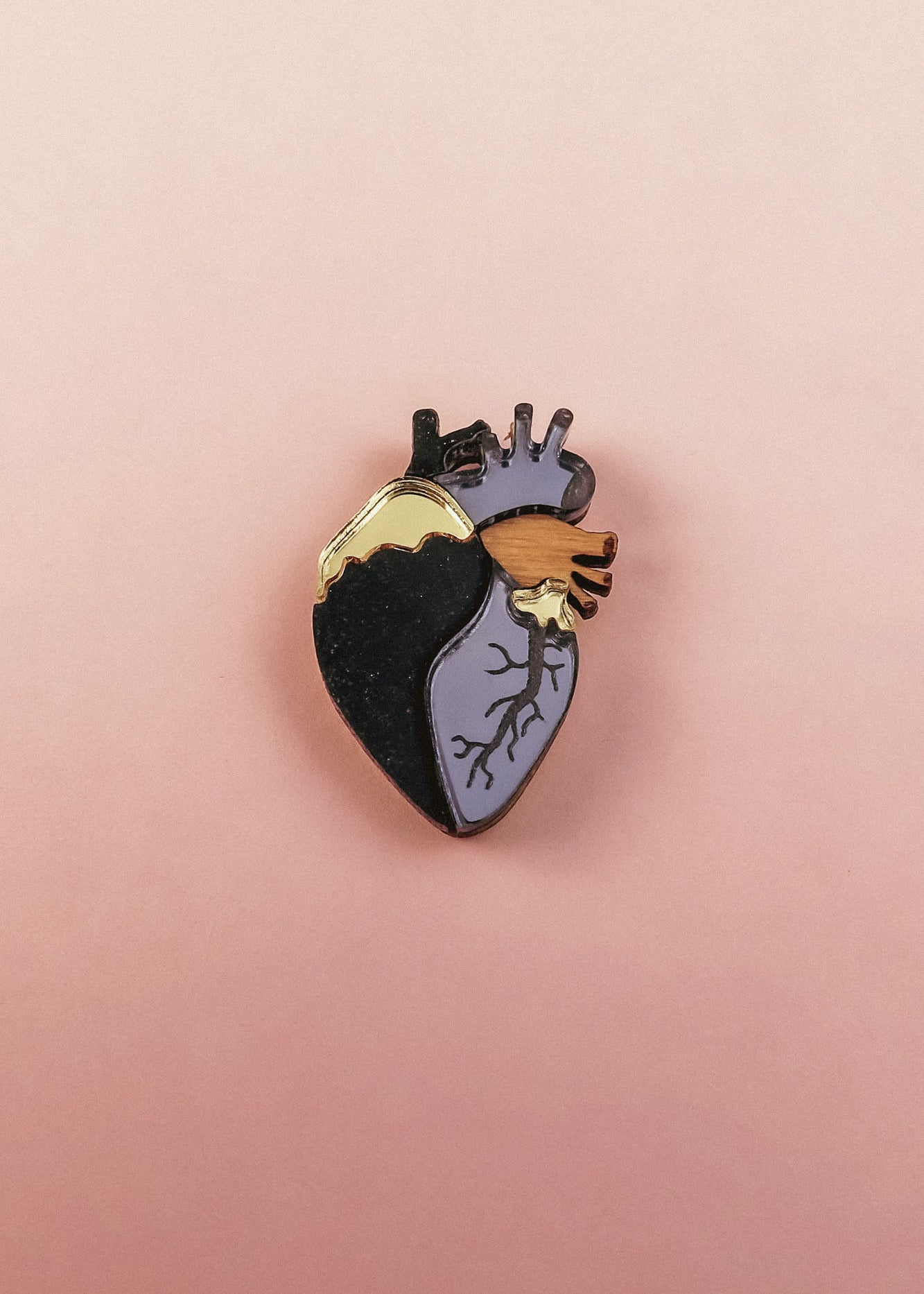 A handcrafted Anatomical Heart Pin with one side black, the other lavender, and highlighted by gold and purple accents. This statement piece rests on a pink background, perfect for adding bold flair to your collection.
