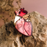 A handcrafted Anatomical Heart Pin, featuring red, pink, and gold details, rests on a textured beige rock with a blurred plant in the background—a true statement piece for any collection.