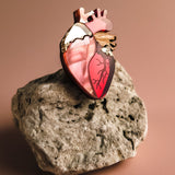 A handcrafted Anatomical Heart Pin, adorned with red and gold accents, serves as a striking statement piece atop a rough, light-colored stone against a soft brown background.