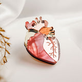 A unique handcrafted Anatomical Heart Pin, shaped with pink, red, gold, and copper hues, is displayed on soft white fabric with blurred dried plants in the foreground.