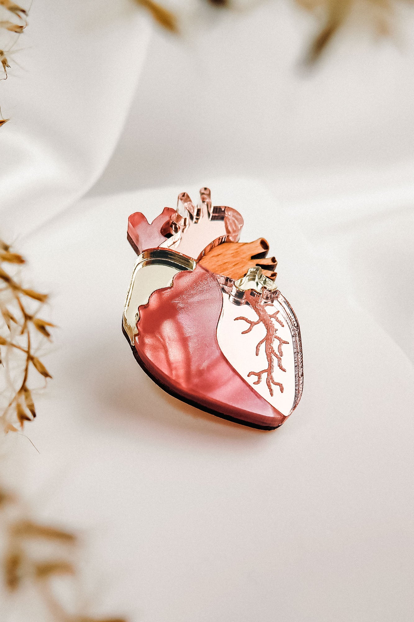 A unique handcrafted Anatomical Heart Pin, shaped with pink, red, gold, and copper hues, is displayed on soft white fabric with blurred dried plants in the foreground.