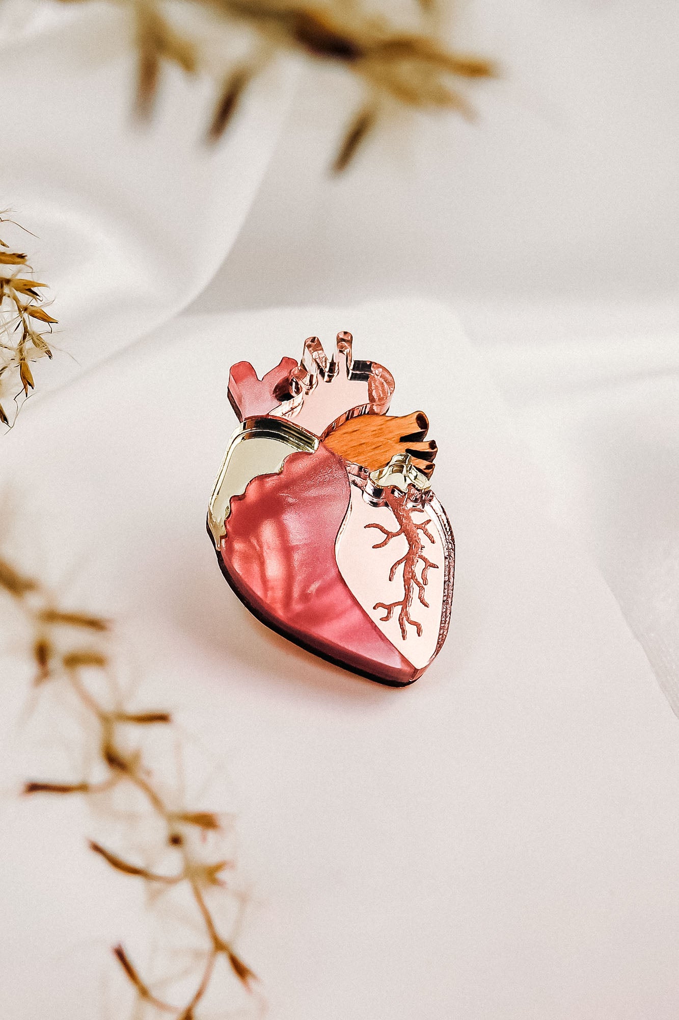 A unique handcrafted glass object shaped like an anatomical heart, featuring shades of pink and gold, sits on a soft white surface with blurred dried plant stems in the foreground.
