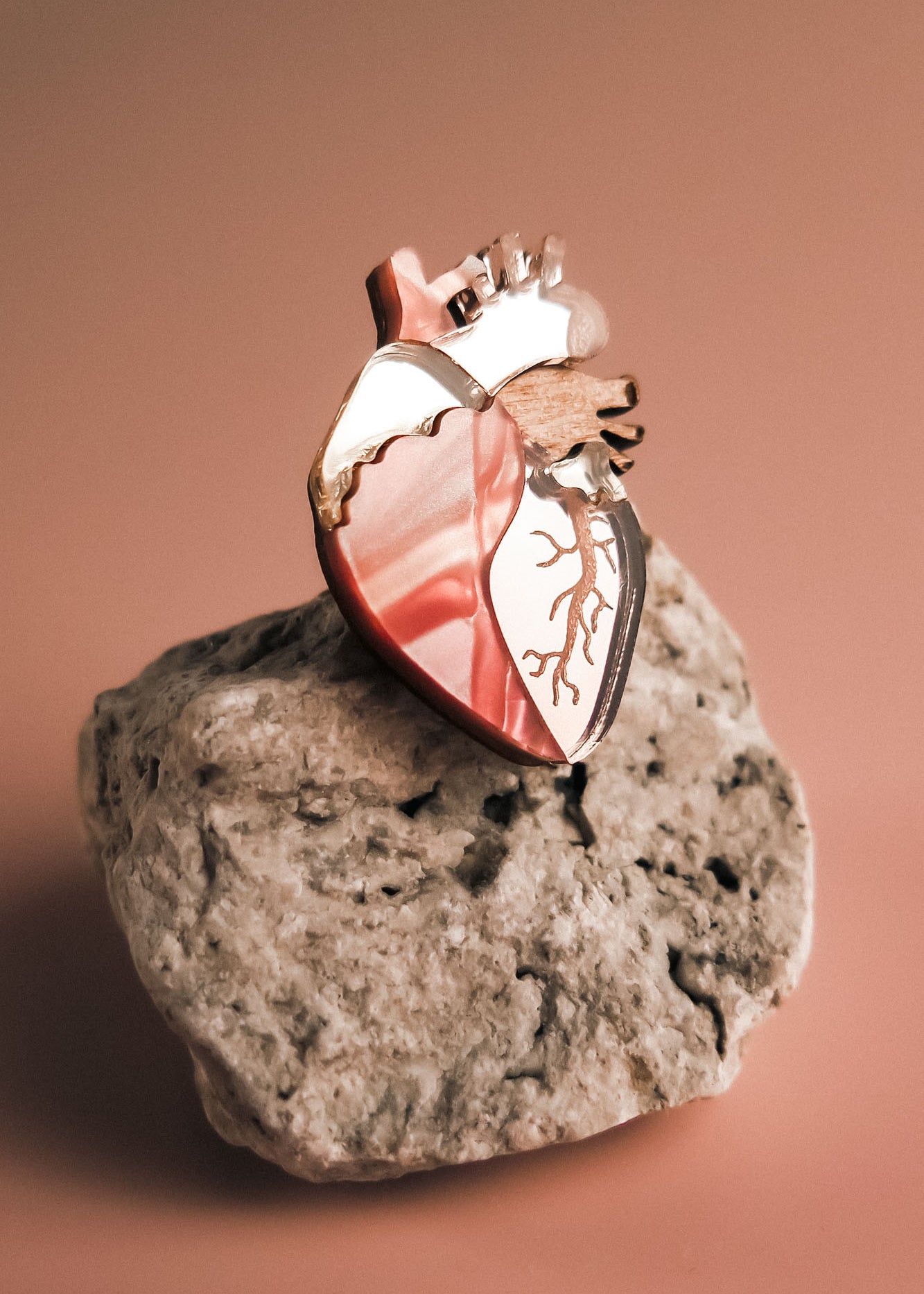 A unique, handcrafted Anatomical Heart Pin in metallic pink rests on a rough, light-colored rock against a soft, warm-toned background.