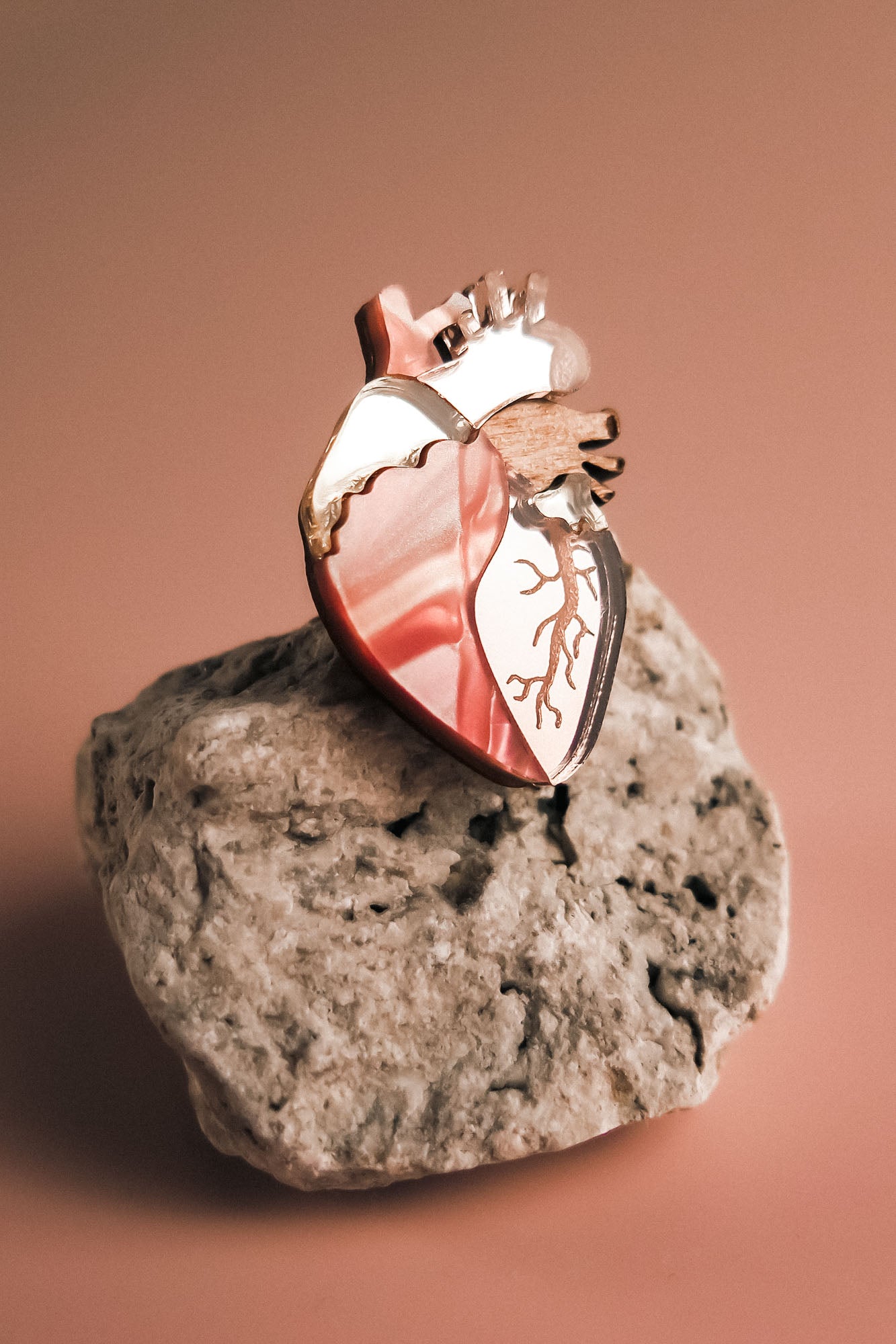 A unique, handcrafted Anatomical Heart Pin in metallic pink rests on a rough, light-colored rock against a soft, warm-toned background.