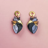 Two Anatomical Heart Earrings with gold, black, and blue details bring a hint of gothic flair, beautifully displayed on a pink background.