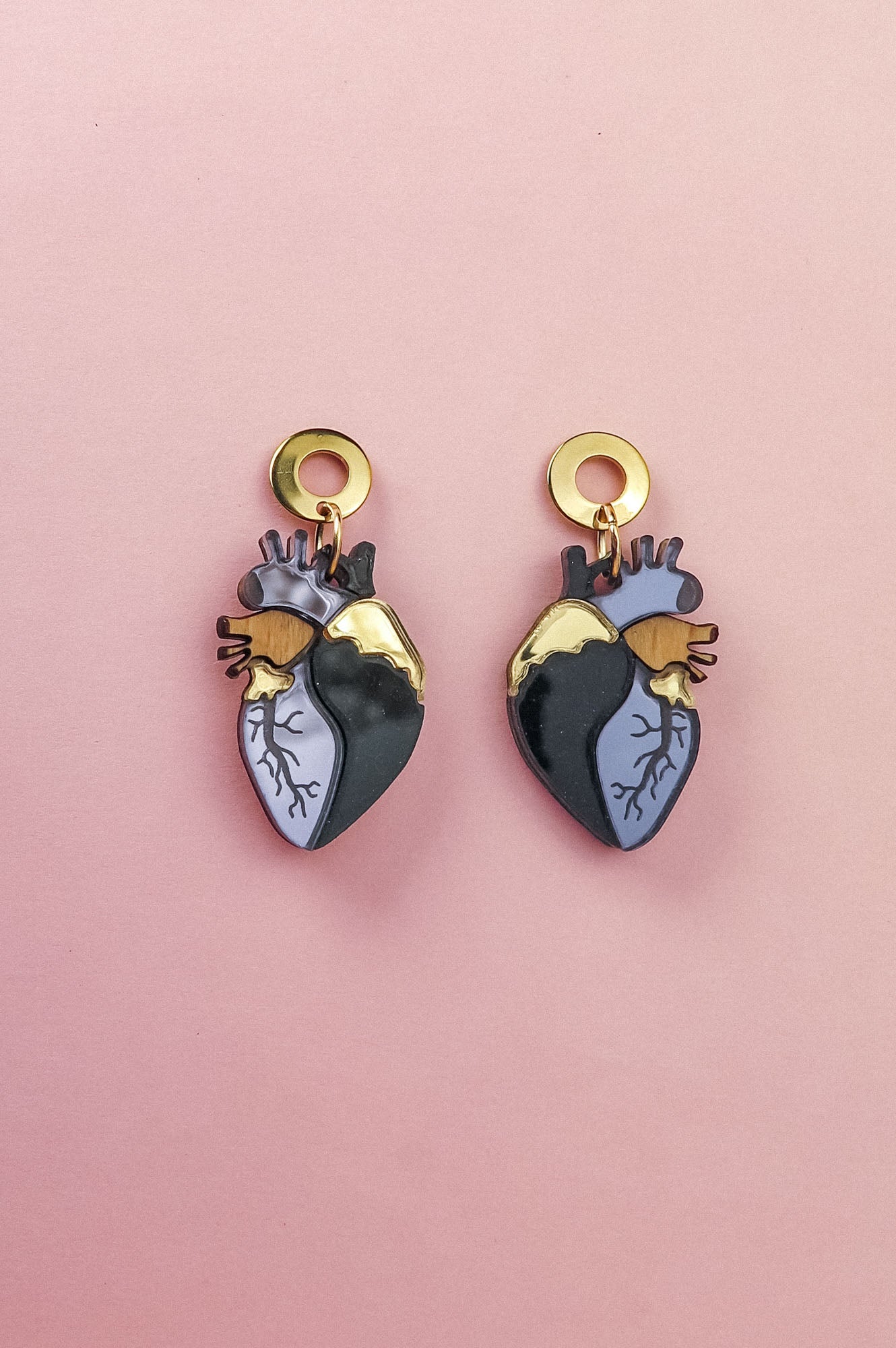 Two Anatomical Heart Earrings with gold, black, and blue details bring a hint of gothic flair, beautifully displayed on a pink background.