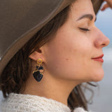 A woman in a brown hat and cream sweater smiles with her eyes closed, showing off handcrafted accessories—a black and gold leafy earring. The background is softly blurred.