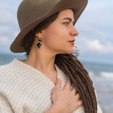 A woman with long brown dreadlocks, wearing a wide-brimmed hat, cream sweater, and handcrafted Anatomical Heart Earrings, stands by the sea with one hand on her chest. She has a tattoo on her forearm and looks serene.