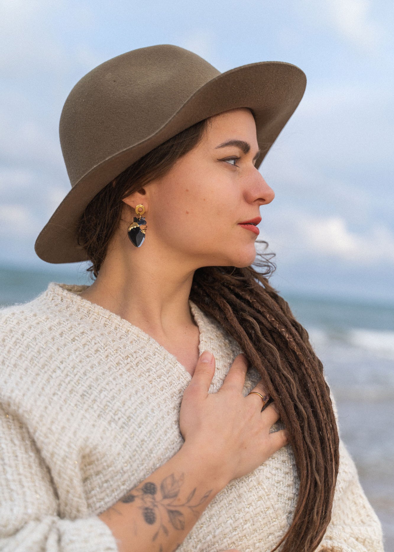 A woman with long brown dreadlocks, wearing a wide-brimmed hat, cream sweater, and handcrafted Anatomical Heart Earrings, stands by the sea with one hand on her chest. She has a tattoo on her forearm and looks serene.