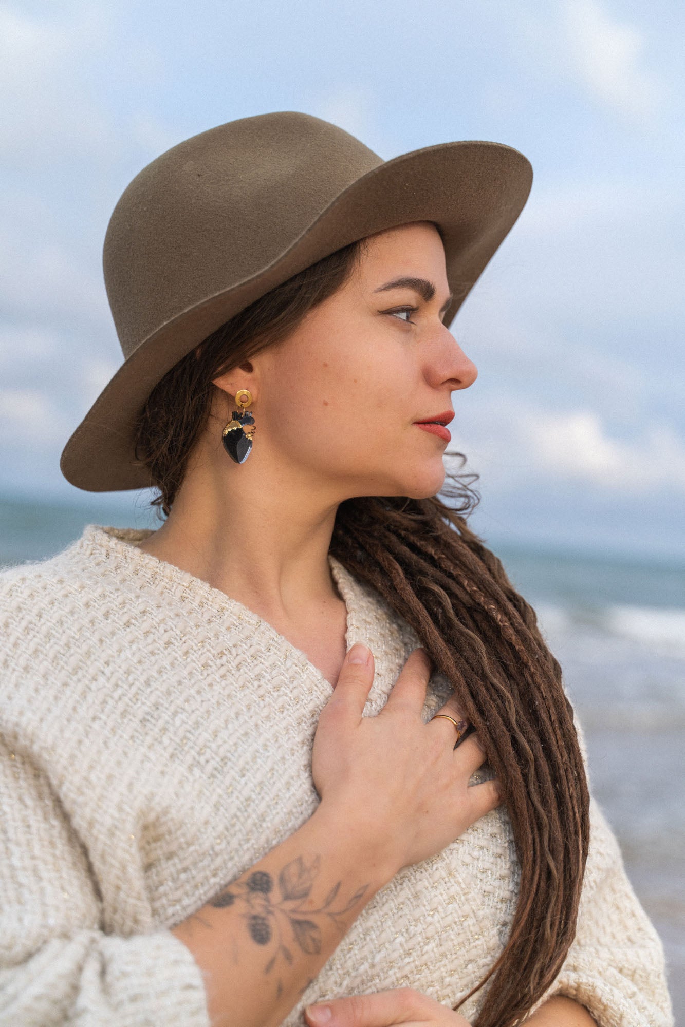 A woman with long brown dreadlocks, wearing a wide-brimmed hat, cream sweater, and handcrafted Anatomical Heart Earrings, stands by the sea with one hand on her chest. She has a tattoo on her forearm and looks serene.