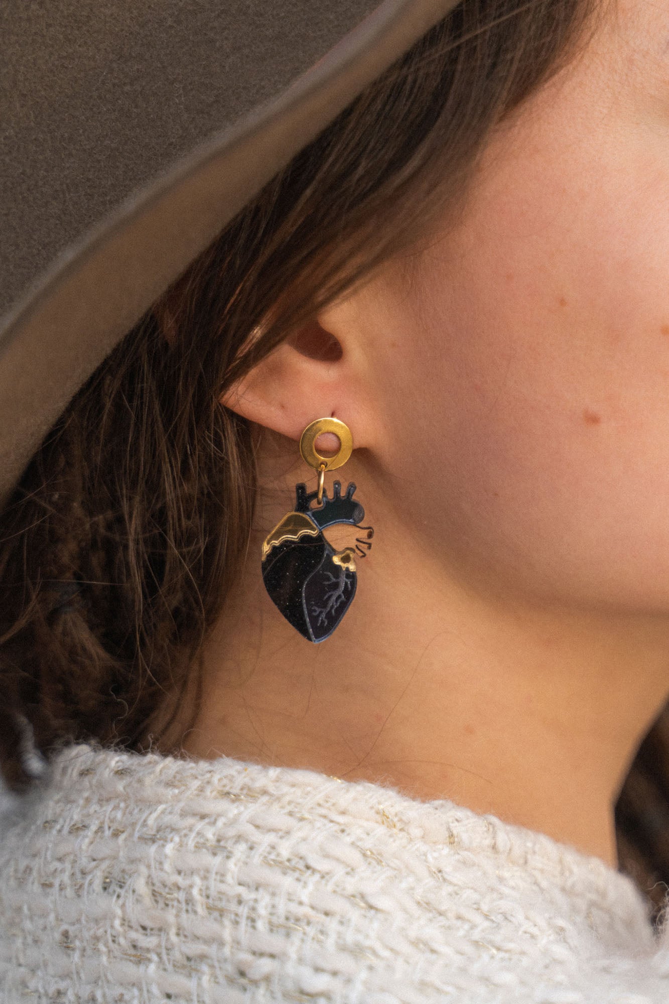 Close-up of a person wearing a gold and black Anatomical Heart Earring with gothic flair. The individual has light skin, brown hair, and is dressed in a beige hat and cream knitted sweater.