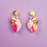 A pair of bold style Anatomical Heart Earrings crafted from holographic acrylic, with gold-toned tops and pink and red details, displayed on a pastel purple background.