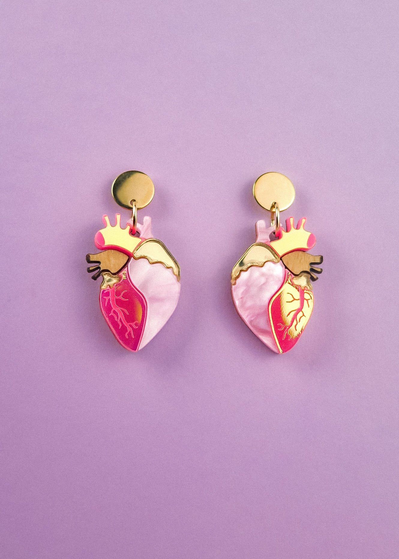 A pair of bold style Anatomical Heart Earrings crafted from holographic acrylic, with gold-toned tops and pink and red details, displayed on a pastel purple background.