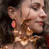 A woman with wavy brown hair and closed eyes stands peacefully among dry brown branches, wearing bold style pink and gold earrings and a red top.