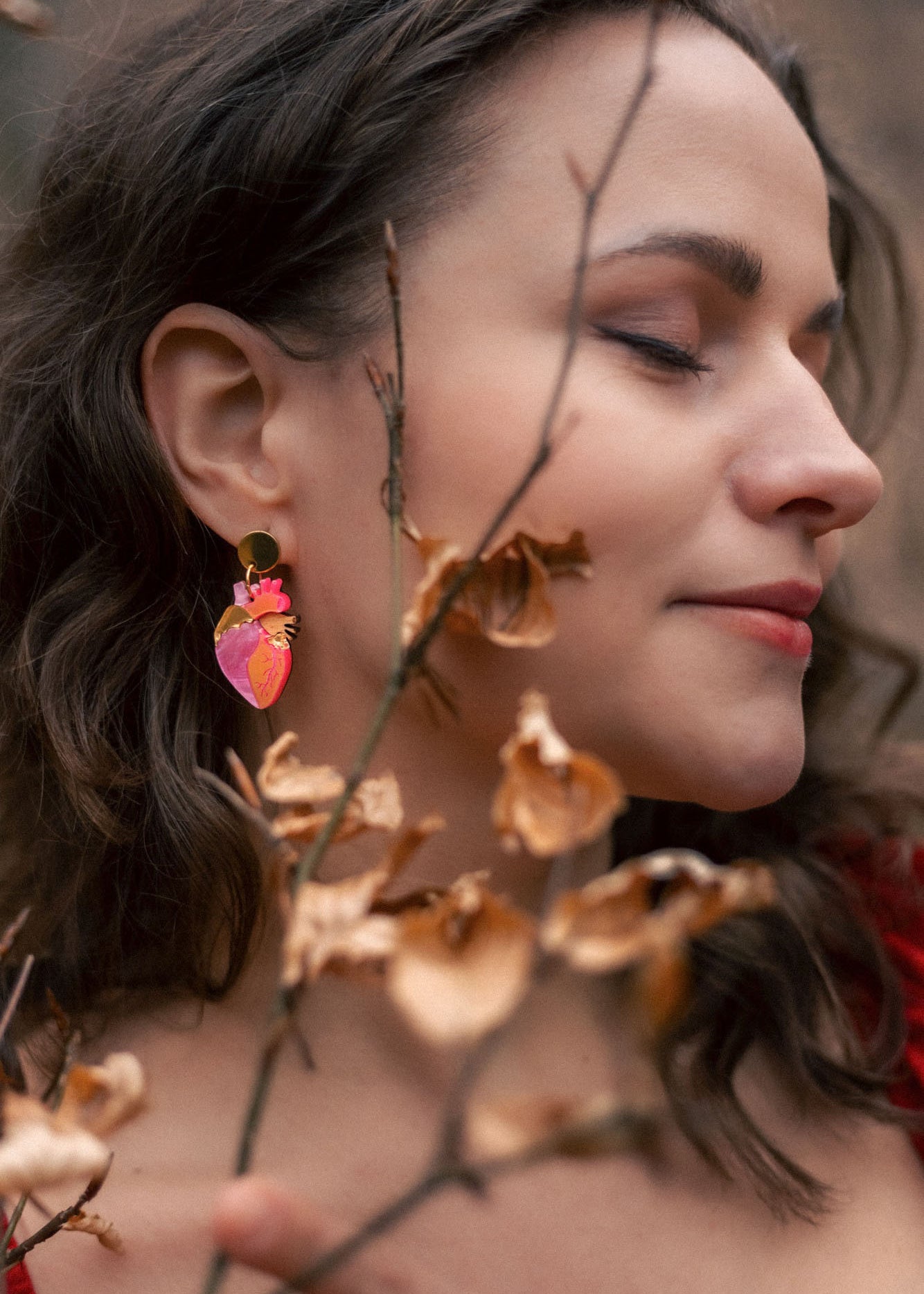 A woman with wavy brown hair and closed eyes stands peacefully among dry brown branches, wearing bold style pink and gold earrings and a red top.