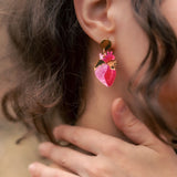 A close-up of a person’s ear wearing bold Anatomical Heart Earrings crafted from pink and gold holographic acrylic. Their curled brown hair partially frames the ear, while their hand gently touches their neck.