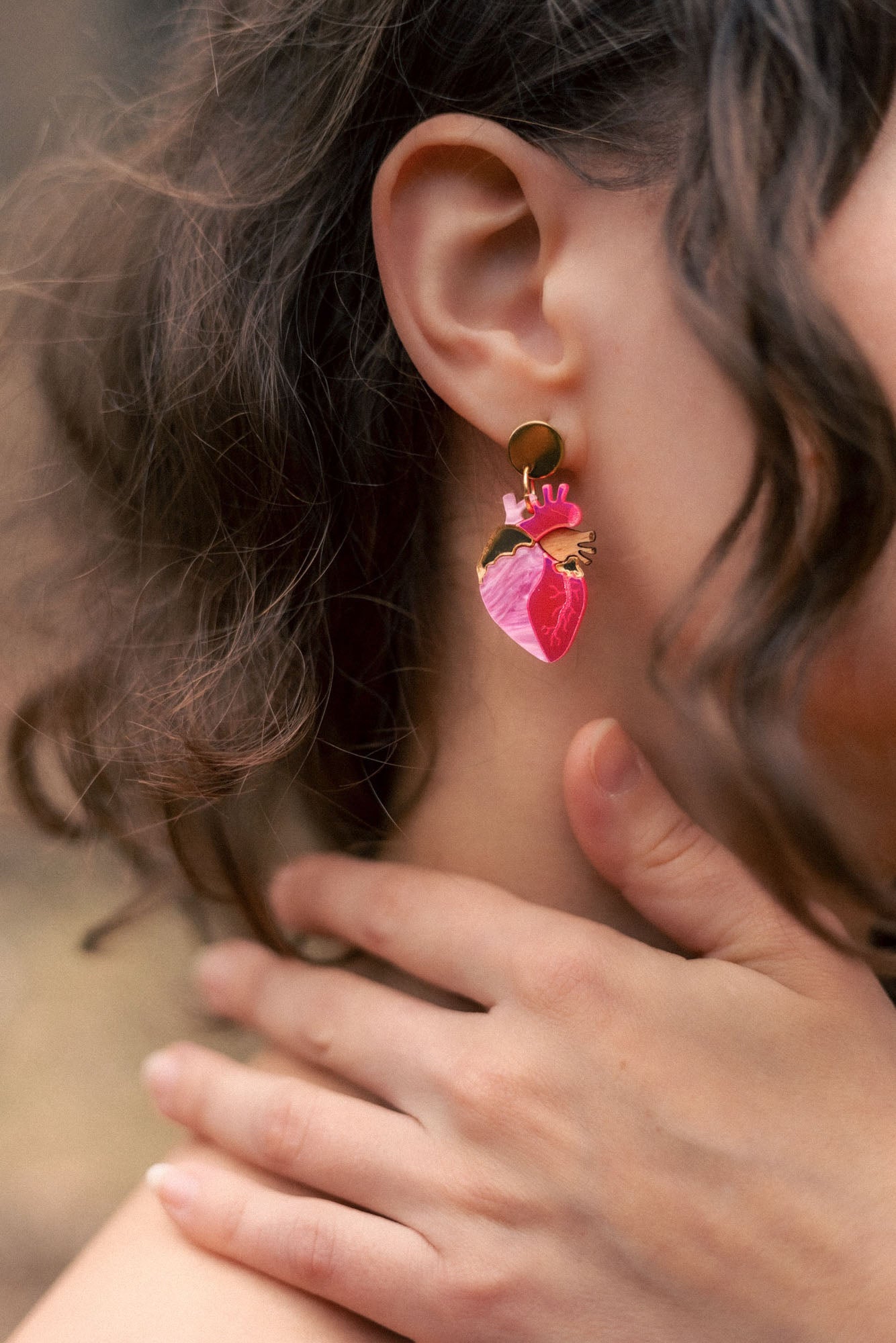 A close-up of a person’s ear wearing bold Anatomical Heart Earrings crafted from pink and gold holographic acrylic. Their curled brown hair partially frames the ear, while their hand gently touches their neck.