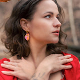 A woman with wavy brown hair and a tattooed forearm crosses her hands over her chest. She wears a red dress, bold style pink statement earrings, and stands outdoors among brown autumn leaves, looking to the side.