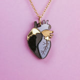 A gold chain anatomical heart necklace with a pendant featuring black, gray, and gold sections is displayed against a pink background—a truly unique jewelry piece.