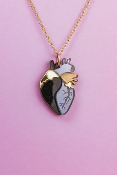 A gold chain anatomical heart necklace with a pendant featuring black, gray, and gold sections is displayed against a pink background—a truly unique jewelry piece.