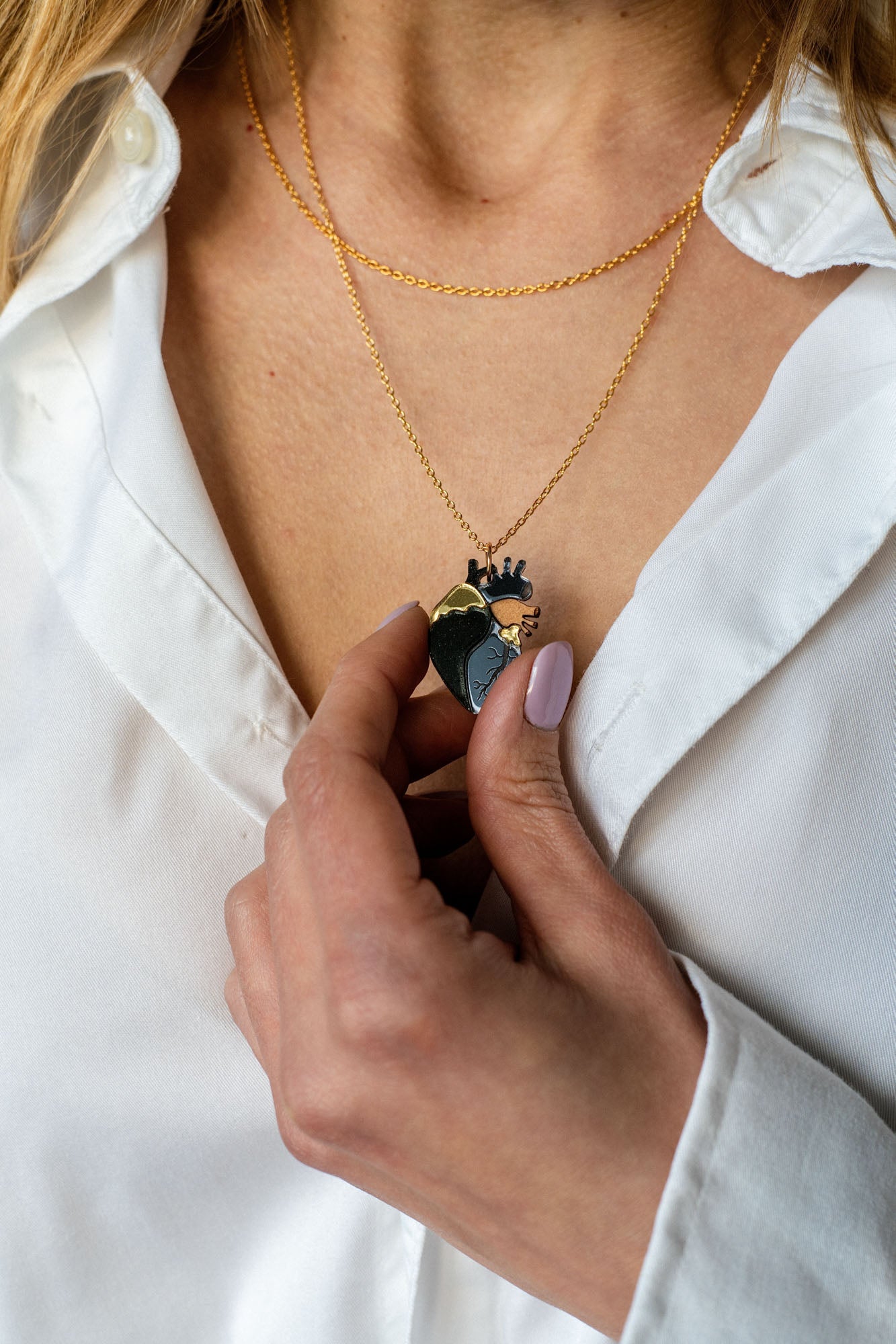 A person wearing a white shirt holds a unique jewelry piece—a gold anatomical heart necklace with a crown-shaped pendant. Their nails are painted a pale pink color, adding elegance to the handcrafted jewelry design.