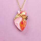 A gold chain anatomical heart necklace, featuring pink, gold, and orange enamel details, is displayed against a light purple background—a truly unique jewelry piece for those who love handcrafted jewelry.