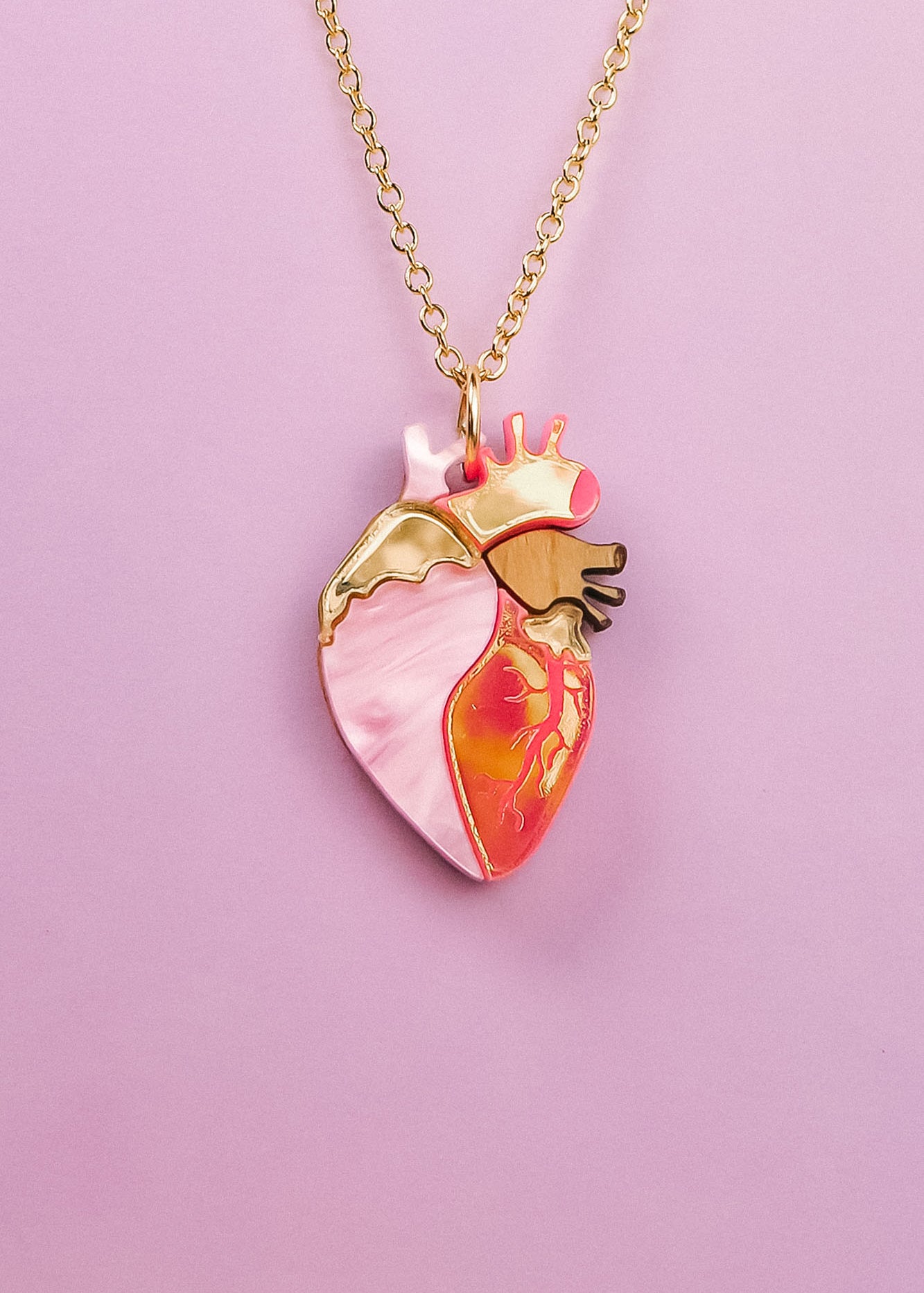 A gold chain anatomical heart necklace, featuring pink, gold, and orange enamel details, is displayed against a light purple background—a truly unique jewelry piece for those who love handcrafted jewelry.
