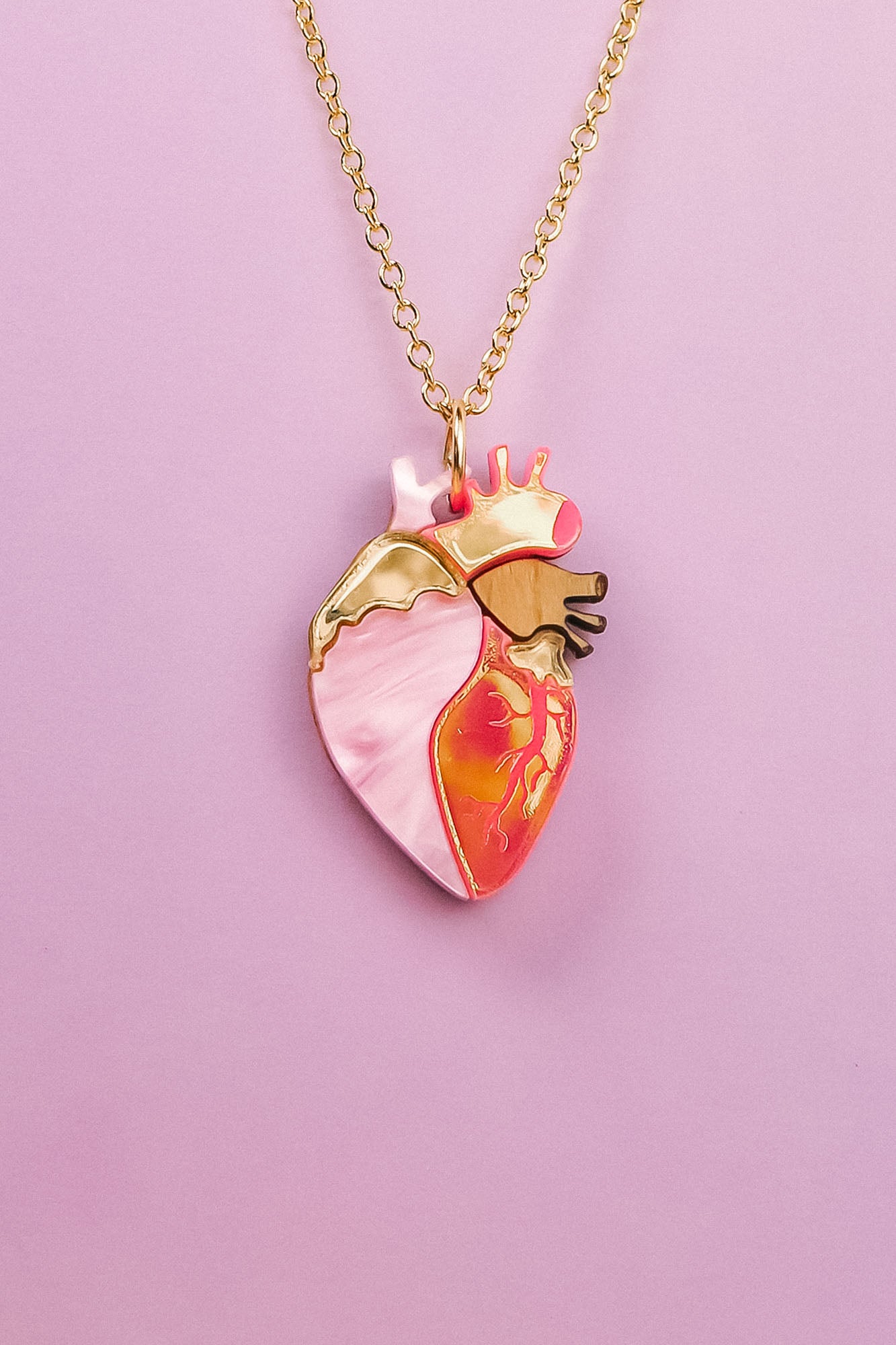 A gold chain anatomical heart necklace, featuring pink, gold, and orange enamel details, is displayed against a light purple background—a truly unique jewelry piece for those who love handcrafted jewelry.