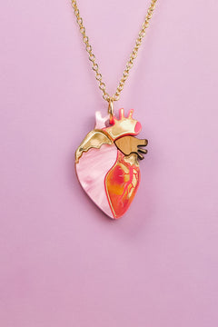 A gold chain anatomical heart necklace, featuring pink, gold, and orange enamel details, is displayed against a light purple background—a truly unique jewelry piece for those who love handcrafted jewelry.