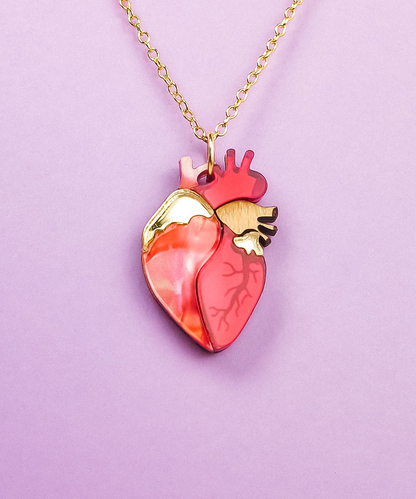 A gold chain anatomical heart necklace in shades of red, pink, and gold, displayed on a light purple background—a unique jewelry piece sure to stand out.
