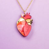 A gold chain anatomical heart necklace in shades of red, pink, and gold, displayed on a light purple background—a unique jewelry piece sure to stand out.