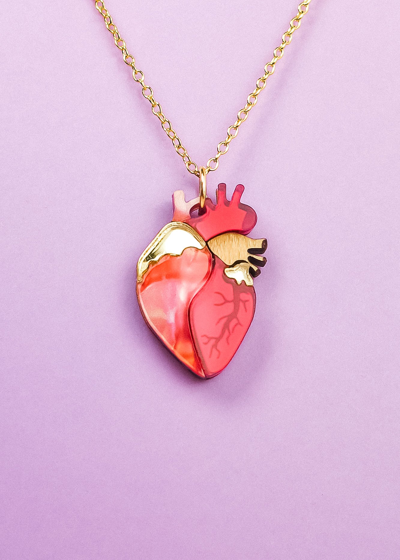 A gold chain anatomical heart necklace in shades of red, pink, and gold, displayed on a light purple background—a unique jewelry piece sure to stand out.