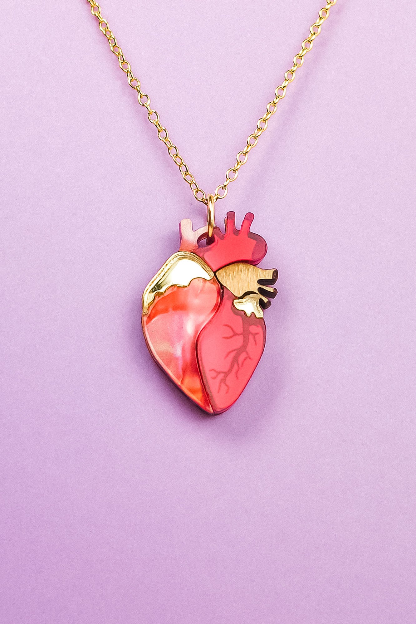 A gold chain anatomical heart necklace in shades of red, pink, and gold, displayed on a light purple background—a unique jewelry piece sure to stand out.