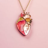 A handcrafted gold chain necklace with a pendant shaped like an anatomical human heart, featuring pink, gold, and cream accents—this unique jewelry piece is beautifully displayed on a pale pink background.