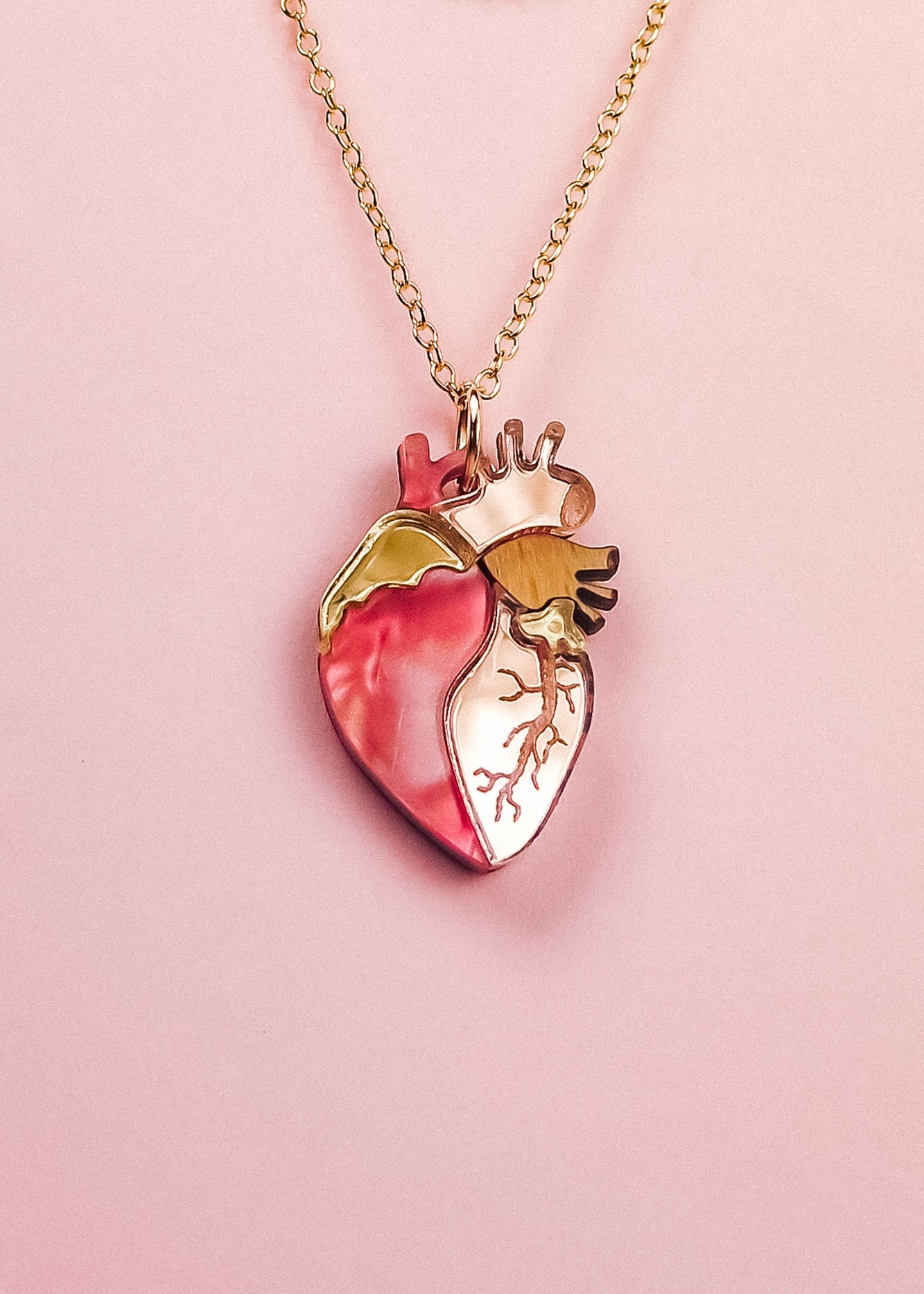 A handcrafted gold chain necklace with a pendant shaped like an anatomical human heart, featuring pink, gold, and cream accents—this unique jewelry piece is beautifully displayed on a pale pink background.
