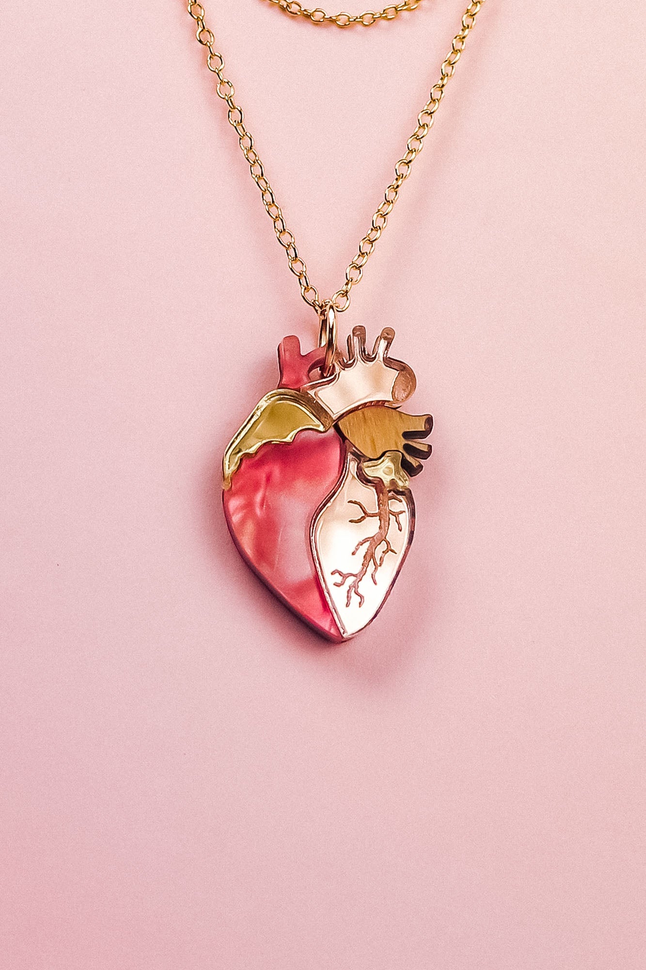 A handcrafted gold chain necklace with a pendant shaped like an anatomical human heart, featuring pink, gold, and cream accents—this unique jewelry piece is beautifully displayed on a pale pink background.
