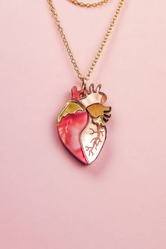 A handcrafted gold chain necklace with a pendant shaped like an anatomical human heart, featuring pink, gold, and cream accents—this unique jewelry piece is beautifully displayed on a pale pink background.