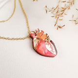 A gold anatomical heart necklace with a pendant shaped like a human heart, featuring pink, gold, and brown detailing, is displayed on a light surface with delicate dried grass in the background—perfect for lovers of unique jewelry.