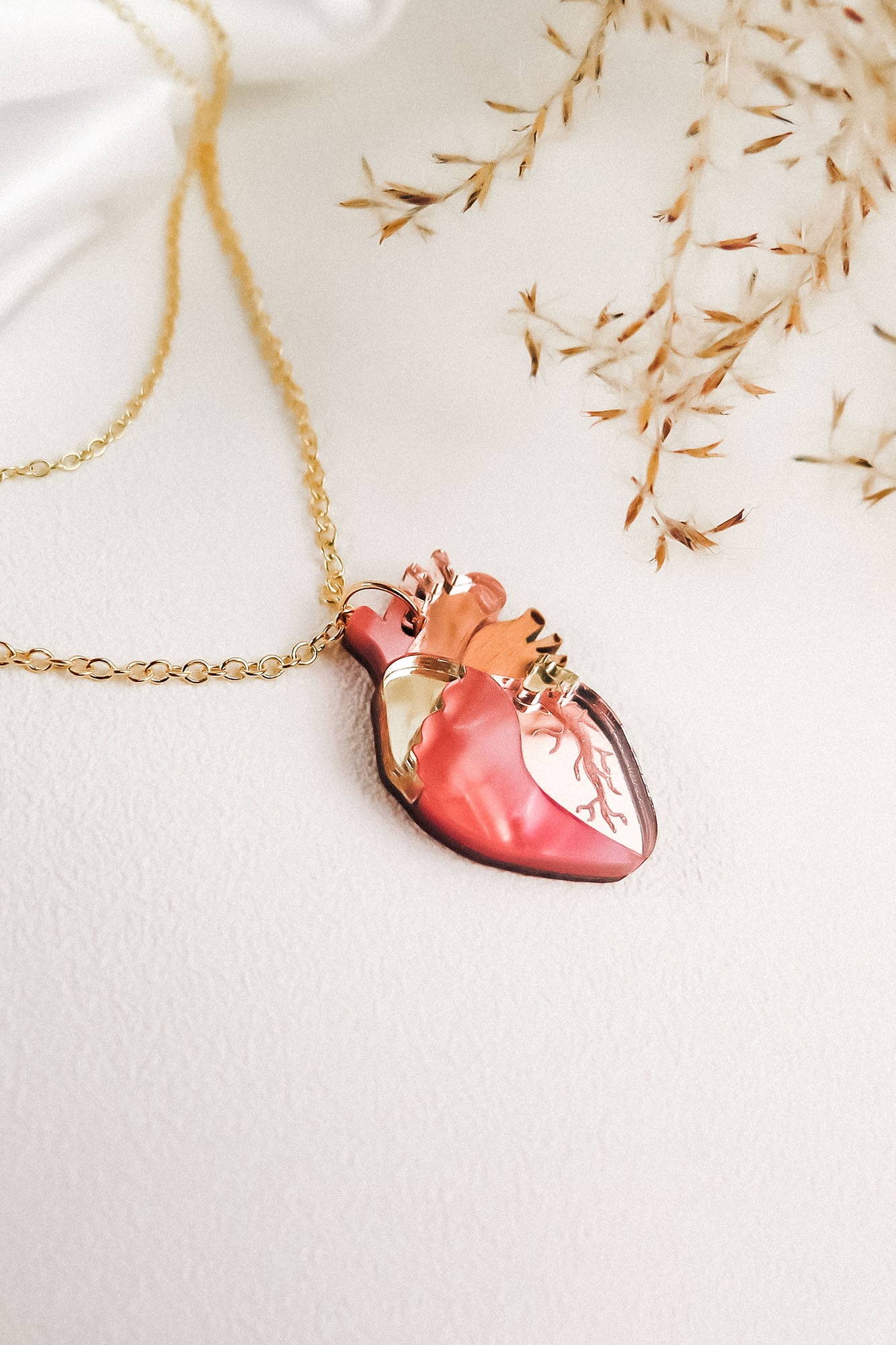 A gold anatomical heart necklace with a pendant shaped like a human heart, featuring pink, gold, and brown detailing, is displayed on a light surface with delicate dried grass in the background—perfect for lovers of unique jewelry.