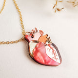 A handcrafted gold chain necklace with a pendant shaped like an anatomical heart, featuring pink, gold, and copper accents, rests on a white surface with dried grasses in the background—a unique jewelry piece.
