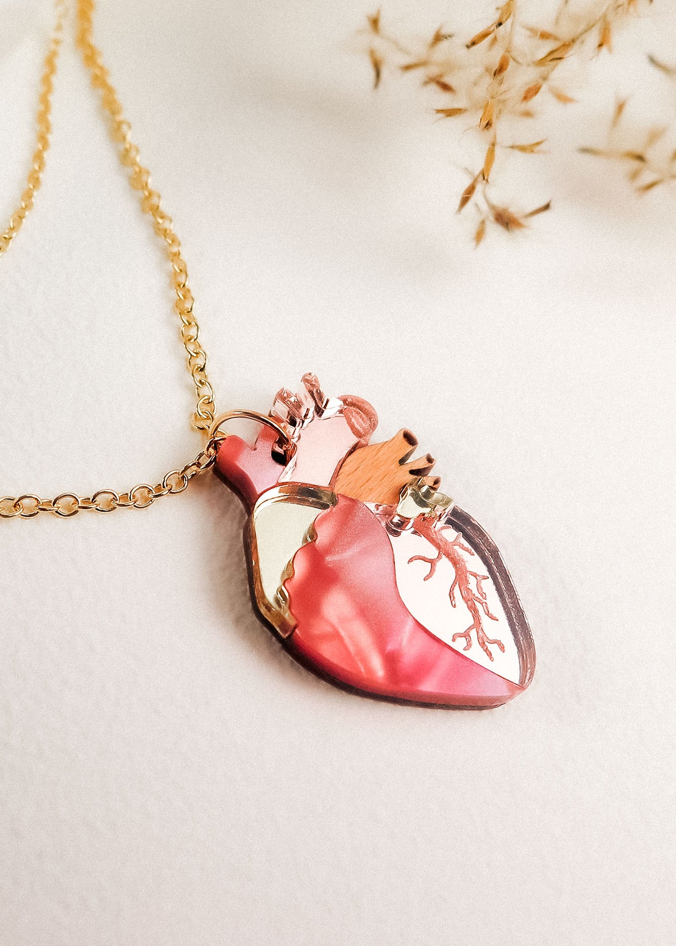 A handcrafted gold chain necklace with a pendant shaped like an anatomical heart, featuring pink, gold, and copper accents, rests on a white surface with dried grasses in the background—a unique jewelry piece.