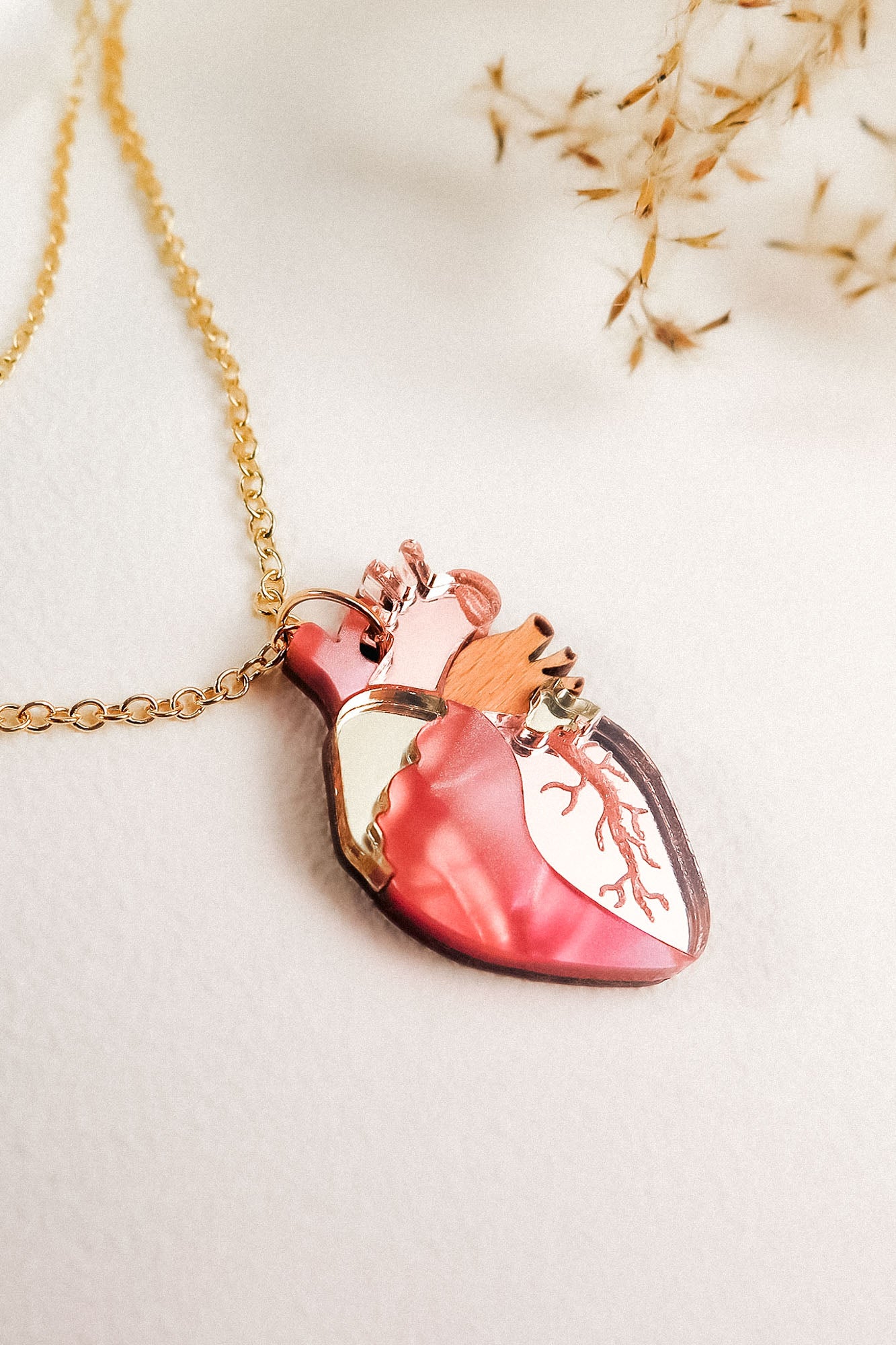 A handcrafted gold chain necklace with a pendant shaped like an anatomical heart, featuring pink, gold, and copper accents, rests on a white surface with dried grasses in the background—a unique jewelry piece.