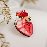 A handcrafted perfume bottle shaped like an anatomical heart, featuring red and pink tones with vein-like details, rests on a soft white surface beside dried grasses—truly a statement piece for any collection.