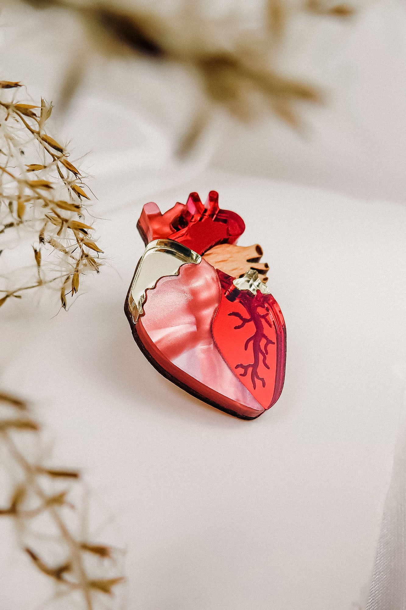 A handcrafted perfume bottle shaped like an anatomical heart, featuring red and pink tones with vein-like details, rests on a soft white surface beside dried grasses—truly a statement piece for any collection.